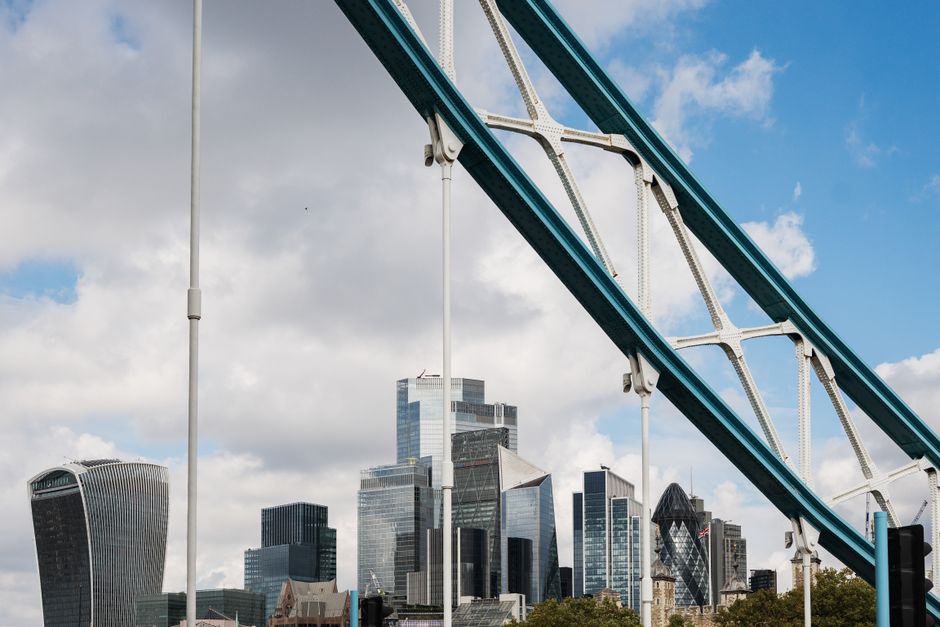 A high-angle view of the Tower Bridge spanning the River Thames in London, with its two towers and suspension structure prominently visible. Behind the bridge, the modern skyline features numerous tal